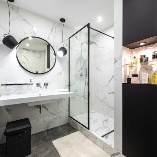 Shower room against washbasin and mirror between shiny lights in bathroom with assorted perfume in bottles on shelves